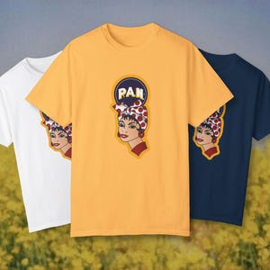 Puede incluir: Tres camisetas en blanco, amarillo y azul marino. Cada camiseta presenta un gráfico de una mujer con un pañuelo de lunares rojos y blancos y la palabra "PAN" en un círculo. Las camisetas se muestran frente a un campo de flores amarillas.