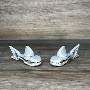 May include: Two white plastic shark-shaped clothespins with teeth and a fin on top.