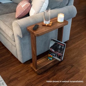 May include: A rectangular walnut side table with a flat top and a lower shelf. The top surface holds a clear glass of water on a wooden coaster, a white candle in a glass jar, and a black remote control. A book and a small cube puzzle sit below.