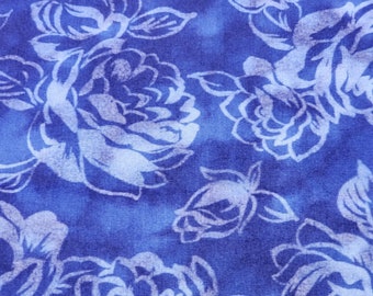 May include: Close-up of a fabric with a repeating floral pattern. The design features large, stylized white roses with detailed petals and leaves against a deep blue background. The fabric has a slightly textured appearance, with variations in the blue hue.