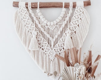 Puede incluir: Un tapiz de macramé blanco con una barra de madera en la parte superior. El macramé tiene un patrón de diamante con flecos en la parte inferior.