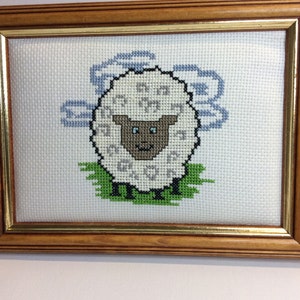 May include: A framed cross-stitch embroidery of a white sheep with a brown face and blue eyes. The sheep is standing on green grass with blue clouds in the background.