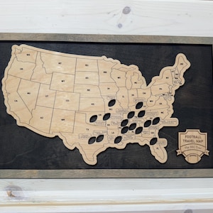 May include: Wooden map of the United States with state outlines and football-shaped cutouts. The map is framed in a gray-toned wood and mounted on a dark background. A small sign reads "FOOTBALL TRAVEL MAP" with a football graphic.