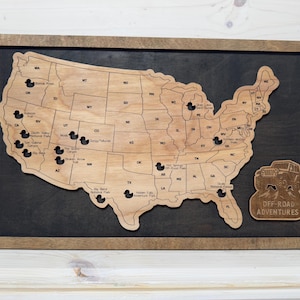 Puede incluir: Mapa de madera de los Estados Unidos con contornos de estados y alfileres en forma de pato que marcan ubicaciones. El mapa está en un fondo oscuro dentro de un marco de madera. Un emblema de madera separado dice "Off-Road Adventures".
