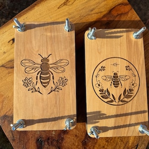 Pode incluir: Dois blocos de impressão de madeira com desenhos de abelhas e flores. Os blocos são de madeira clara e têm braçadeiras de metal nas laterais. Os desenhos de abelhas são gravados na madeira.