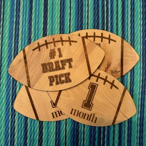Puede incluir: Cuatro letreros de madera con forma de balón de fútbol con texto grabado. El letrero superior dice "#1 DRAFT PICK". Los otros tres letreros dicen "1 mes", "2 meses" y "3 meses".