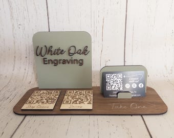 Op de afbeelding: Een houten visitekaartjeshouder met een groen bord waarop "White Oak Engraving" staat. Er zijn twee QR-codekaartjes met de tekst "Shop Online" en "Socials" en een visitekaartje met een QR-code en contactgegevens. Het bord zegt "Take One".