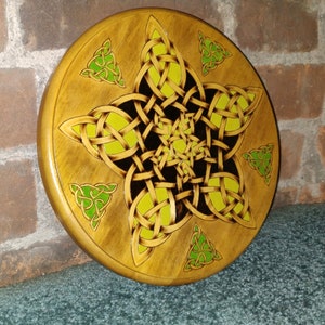 May include: A round wooden plaque with a Celtic knot design in shades of yellow, green, and black. The knot is in the shape of a star with five points.