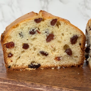 May include: A slice of golden-brown raisin bread, showcasing a light, airy interior dotted with dark raisins. The bread sits on a wooden cutting board, with a marble background.