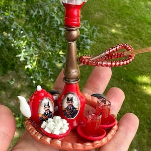 May include: A miniature red and gold hookah set with a tray, teapot, two glasses, and sugar cubes. The hookah has a red base and a gold stem with a silver top. The teapot and glasses are also red with gold accents. The tray is brown with a gold rim.