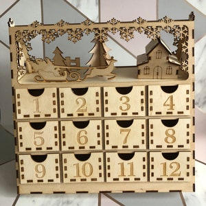 Könnte beinhalten: Holz-Adventskalender mit 12 Schubladen, die jeweils von 1 bis 12 nummeriert sind. Die Oberseite des Kalenders zeigt ein ausgeschnittenes Design einer Schneelandschaft mit einem Haus, einem Weihnachtsbaum und einem Rentier, das einen Schlitten zieht.