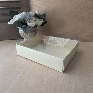May include: A rectangular, off-white gift box with a clear plastic lid. The box is empty and sits on a light-colored wooden surface. A decorative vase with white and green flowers is in the background.