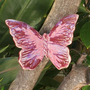 May include: A ceramic butterfly sculpture with a glossy, textured finish. The butterfly is primarily burgundy with lighter pink and white accents. The wings are detailed with etched patterns, and the sculpture is mounted on a tree branch.