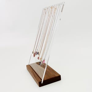 May include: A wooden and clear acrylic jewelry display stand holding several necklaces with various pendant designs. The stand has a rectangular wooden base and a clear acrylic panel with slots to hang the necklaces.