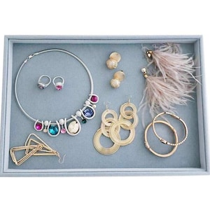 May include: A light blue velvet jewelry tray with compartments for storing necklaces, earrings, and rings. The tray features a silver necklace with colorful gemstones, gold hoop earrings, and a pair of feather earrings.