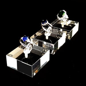 May include: Three clear acrylic ring display stands with three rings on them. The rings are silver with different colored gemstones. The first ring has a green gemstone, the second ring has a blue gemstone, and the third ring has a blue gemstone.