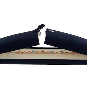 May include: A navy blue jewelry box with a cream-colored interior, open to reveal a gold-toned bracelet with colorful gemstones. The box has a hinged design and a tassel closure.