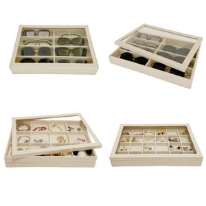 May include: Beige sunglasses and jewelry organizers with clear lids. The organizers have multiple compartments for storing sunglasses, necklaces, bracelets, earrings, and rings. The organizers are made of a soft, neutral-colored material.