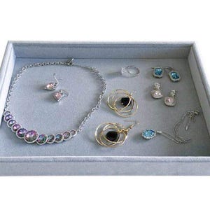 May include: A gray jewelry tray filled with various pieces of jewelry. The jewelry includes a silver necklace with iridescent stones, earrings, rings, and gold-toned earrings with black stones. Some pieces have blue or clear stones.