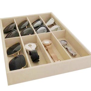 May include: Beige jewelry organizer with multiple compartments. Contains sunglasses and watches. The sunglasses have black frames and dark lenses. The watches have silver, gold, and rose gold tones. The organizer is rectangular.