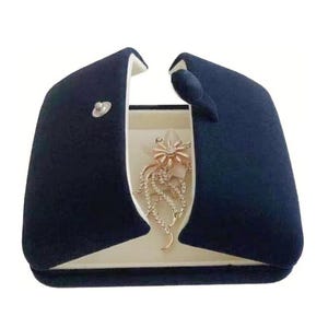 May include: A dark blue velvet jewelry box with a hinged lid. The box is open and a gold necklace with a flower pendant is inside.