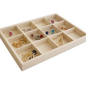 May include: Beige jewelry organizer with multiple compartments, filled with rings, necklaces, and earrings. The organizer is rectangular and has a soft, fabric-like texture. The jewelry includes gold and silver pieces with various gemstones.