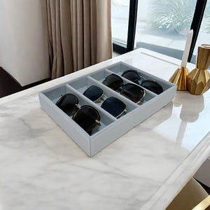 May include: A gray velvet-lined tray with four compartments for storing sunglasses. The tray is on a white marble surface.