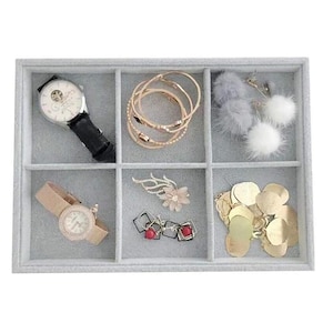 May include: A rectangular gray velvet jewelry organizer tray with six compartments. It contains a black leather watch, a rose gold watch, gold-tone bangles, white and gray pom-pom earrings, a floral brooch, square earrings, and gold-tone circular charms.