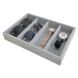 May include: A gray, rectangular jewelry organizer with four compartments. Two pairs of sunglasses, a rose gold watch, and a black watch are inside. The organizer is made of a soft, felt-like material.