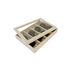May include: A beige rectangular box with a clear lid, designed to store sunglasses. The box has six compartments for organizing sunglasses.