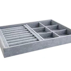 May include: A rectangular, light gray velvet jewelry tray with multiple compartments. The tray features a section with ring slots and several square compartments for organizing jewelry. The soft, plush material provides a protective storage solution.