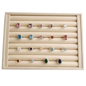 May include: A display tray filled with various gemstone rings. The rings feature different colored stones, including pink, blue, and green, set in gold or silver bands. The tray has a beige, velvet-like surface with multiple rows to hold the rings.