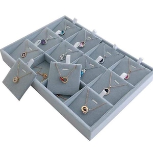 May include: A display tray filled with various necklaces. The necklaces are presented on individual gray velvet cards within a divided tray. The necklaces feature different pendant designs and colors, including gold, silver, and gemstones. The tray is light gray.