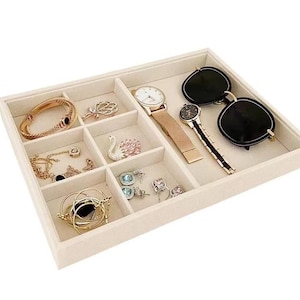 May include: A beige jewelry organizer filled with various accessories. Items include a gold bracelet, necklaces, rings, a swan figurine, two watches, and black sunglasses. The organizer has multiple compartments to store jewelry and accessories.