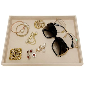 May include: A beige jewelry tray filled with various accessories. Items include gold-toned bracelets, earrings with pearls and red stones, a double G logo, sunglasses, and eyeglasses. The tray is rectangular.