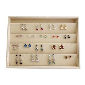 May include: A beige jewelry display case filled with various earrings. The earrings feature different designs, including studs, dangles, and hoops, with gemstones and decorative elements. The case has multiple tiered levels.