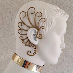 May include: Gold ear cuff with a delicate, intricate design. The cuff is made of metal and has a fan-like shape. It is worn on the upper part of the ear.