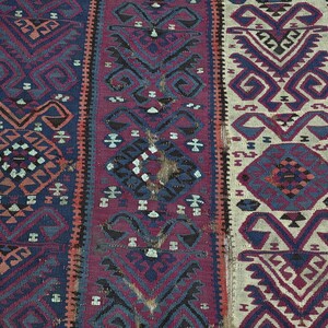 May include: A woven rug featuring vertical panels in deep blue, magenta, and cream. The textile displays repeating geometric patterns, including diamonds and stylized hooked motifs in contrasting shades of dark brown, black, and muted orange.