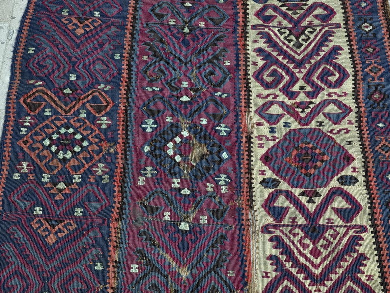 May include: A woven rug featuring vertical panels in deep blue, magenta, and cream. The textile displays repeating geometric patterns, including diamonds and stylized hooked motifs in contrasting shades of dark brown, black, and muted orange.