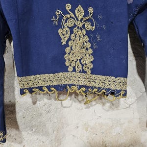 K&ouml;nnte beinhalten: Nahaufnahme eines marineblauen Kleidungsst&uuml;cks mit goldener Stickerei. Das Kleidungsst&uuml;ck hat aufwendige goldene Blumenmuster auf der Vorderseite und entlang der Kanten der &Auml;rmel und des Saums. Der Stoff scheint gealtert zu sein, mit einigen Gebrauchsspuren.