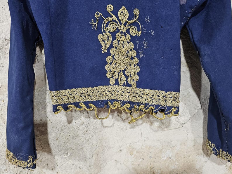 K&ouml;nnte beinhalten: Nahaufnahme eines marineblauen Kleidungsst&uuml;cks mit goldener Stickerei. Das Kleidungsst&uuml;ck hat aufwendige goldene Blumenmuster auf der Vorderseite und entlang der Kanten der &Auml;rmel und des Saums. Der Stoff scheint gealtert zu sein, mit einigen Gebrauchsspuren.
