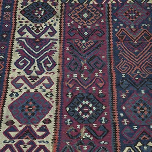 May include: A rectangular woven rug featuring vertical panels with repeating geometric patterns in shades of deep blue, burgundy, cream, and black. The design includes stylized diamond shapes and intricate hooked motifs arranged in a symmetrical layout.