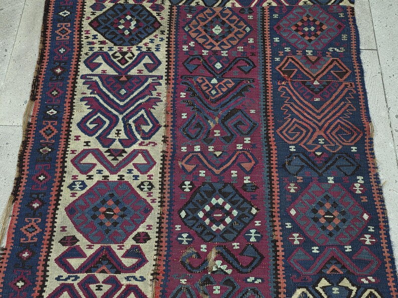 May include: A rectangular woven rug featuring vertical panels with repeating geometric patterns in shades of deep blue, burgundy, cream, and black. The design includes stylized diamond shapes and intricate hooked motifs arranged in a symmetrical layout.