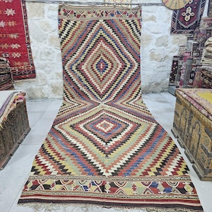 Puede incluir: Una alfombra grande, colorida y tejida a mano con un patrón geométrico de diamantes en tonos rojos, azules, amarillos y marrones. La alfombra tiene un borde de flecos y se muestra en un suelo blanco.