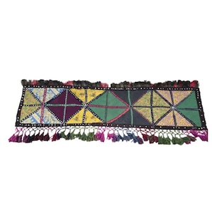 May include: A rectangular table runner featuring a patchwork design of various fabric panels in shades of green, yellow, purple, and blue. The runner is bordered by black fabric with small circular accents and finished with multi-colored tassels.