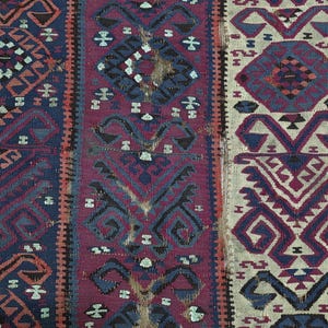 May include: A woven textile featuring vertical panels with intricate geometric patterns in shades of deep blue, magenta, and cream. The design includes repeating diamond and hooked motifs, with visible signs of wear and fraying along the edges.