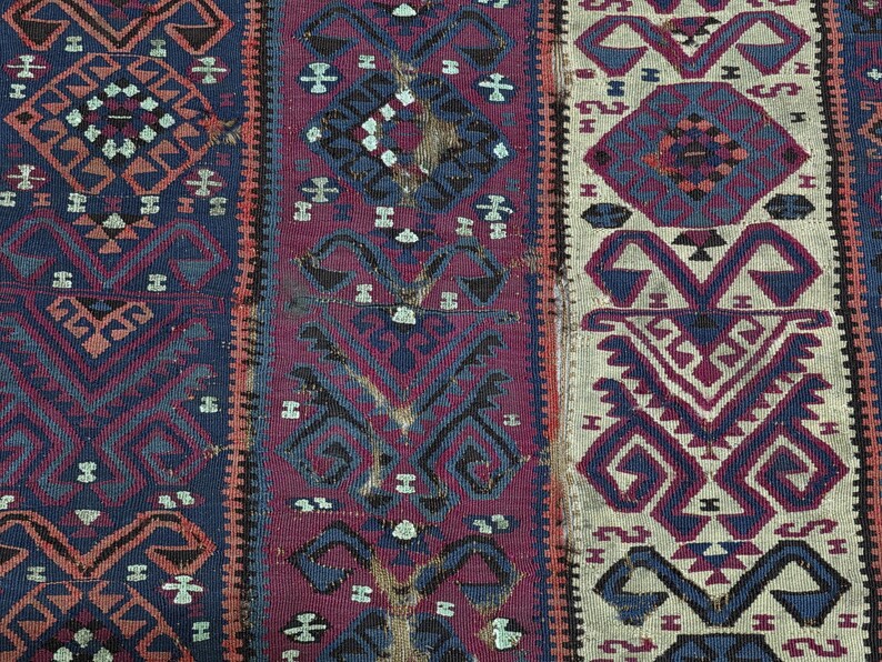 May include: A woven textile featuring vertical panels with intricate geometric patterns in shades of deep blue, magenta, and cream. The design includes repeating diamond and hooked motifs, with visible signs of wear and fraying along the edges.
