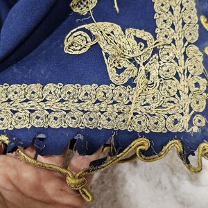 K&ouml;nnte beinhalten: Nahaufnahme eines Teils eines dunkelblauen Stoffes mit aufwendiger goldener Stickerei. Die Stickerei zeigt florale und geometrische Muster an den R&auml;ndern. Der Stoff hat einen gewellten Rand mit goldenem Besatz.