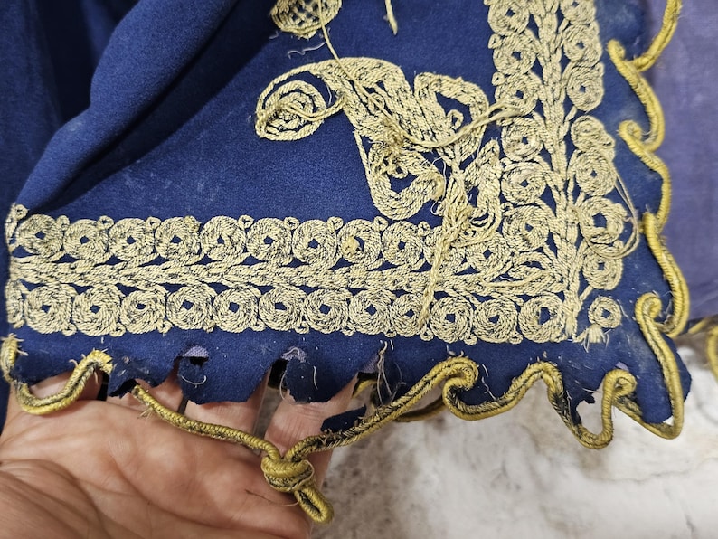 K&ouml;nnte beinhalten: Nahaufnahme eines Teils eines dunkelblauen Stoffes mit aufwendiger goldener Stickerei. Die Stickerei zeigt florale und geometrische Muster an den R&auml;ndern. Der Stoff hat einen gewellten Rand mit goldenem Besatz.