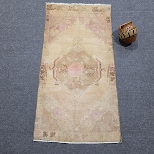 May include: A rectangular area rug featuring a faded beige and tan field with a central pink floral medallion and geometric border patterns, accompanied by a small brown ceramic vessel with black and yellow decorative horizontal bands.